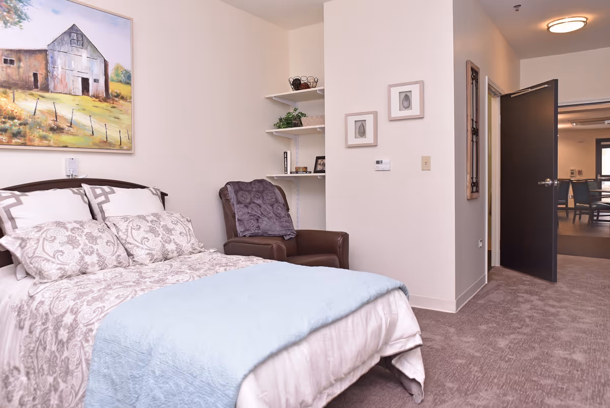 A bedroom with a neatly made bed featuring patterned pillows and a light blue blanket. There is a brown armchair with a purple throw blanket next to the bed. On the wall above the bed is a painting of a barn and a fence in a rural setting. The room has light-colored walls, carpeted floor, and an open door leading to another room with chairs and tables.