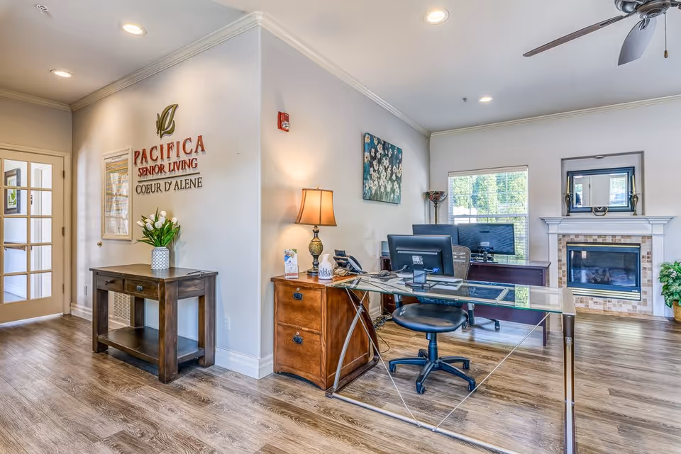 A bright and welcoming office area in a senior living facility with a glass desk, office chair, computer monitors, a wooden side table with a lamp, and a fireplace with a mirror above it. The walls are light-colored, and there is a decorative painting and a vase with flowers on a small wooden table near the entrance. The floor is wood, and there is a ceiling fan above.