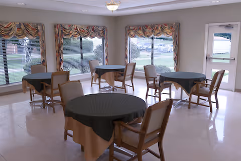 A bright dining room with multiple round tables covered with dark tablecloths and surrounded by wooden chairs with cushioned seats. Large windows with patterned valances let in natural light and offer a view of the outdoor area. A door with a window is visible on the right side of the room.
