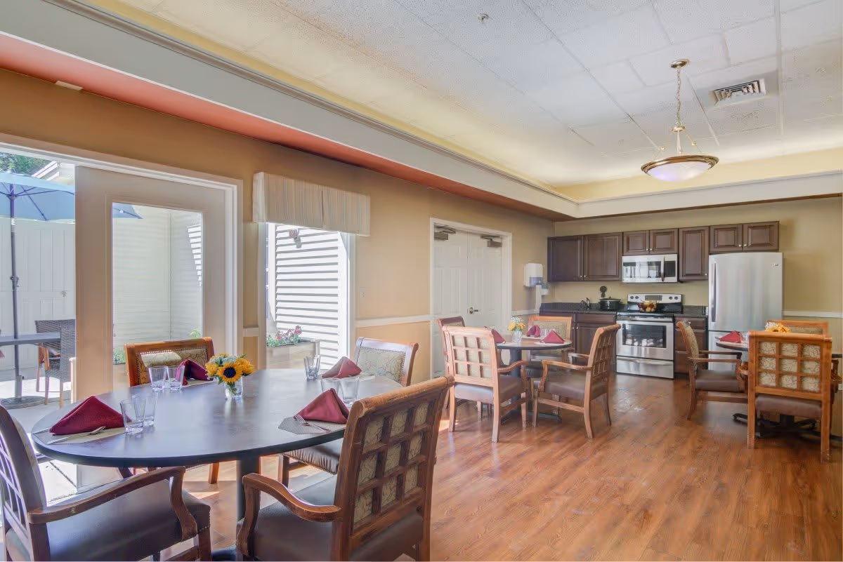 A bright dining area with wooden floors and multiple tables set with glasses and folded burgundy napkins. The room features a kitchen area with stainless steel appliances including a refrigerator, stove, and microwave. There are large windows and a door leading to an outdoor patio with an umbrella and chairs.
