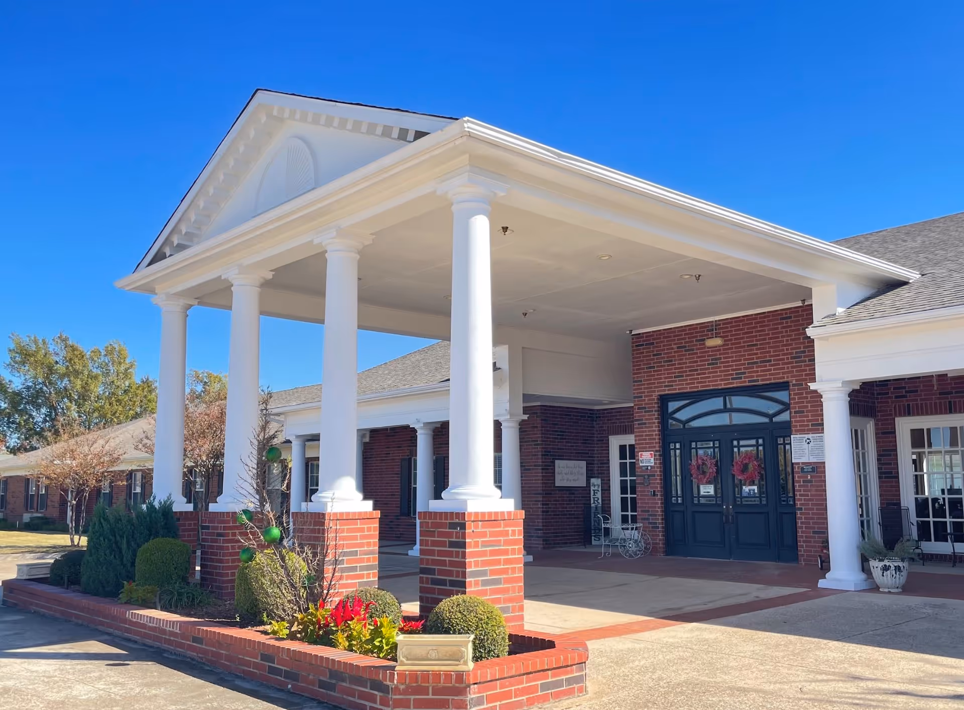 Front entrance of Silver Creek Assisted Living facility featuring a covered portico with white columns, brick walls, double glass doors decorated with wreaths, and landscaped bushes and flowers under a clear blue sky.