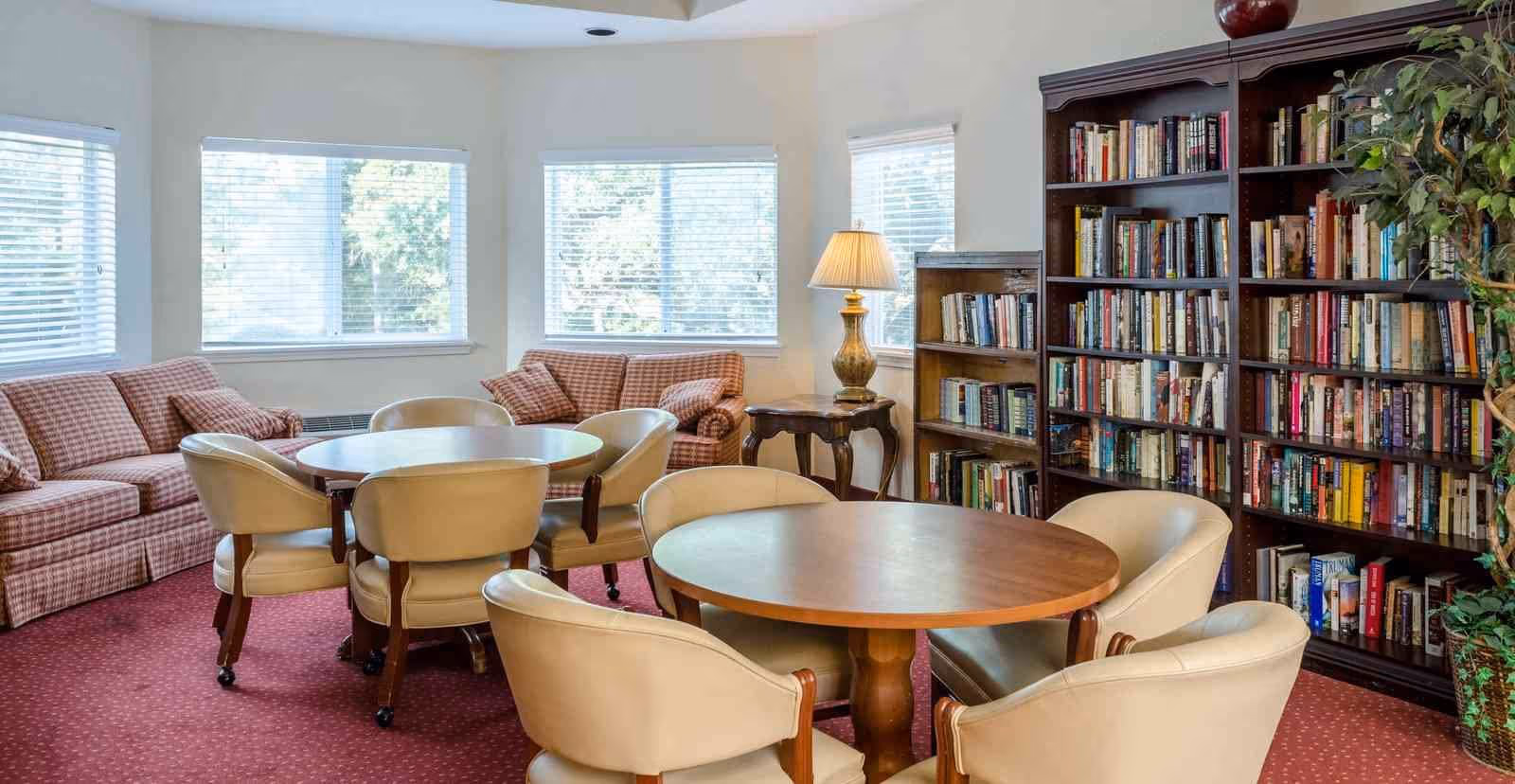 A cozy common area with two round wooden tables surrounded by beige cushioned chairs on a red carpet. There are two plaid sofas near three large windows with white blinds. A wooden bookshelf filled with books and a table lamp on a small side table are also visible.