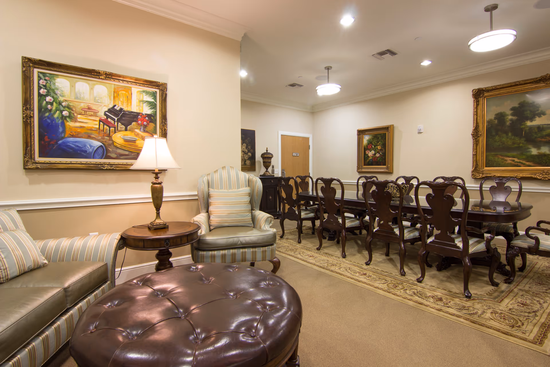 A cozy interior room featuring a seating area with a striped armchair, a matching sofa, a round wooden side table with a lamp, and a large tufted leather ottoman. In the background, there is a long wooden dining table with multiple ornate chairs on a patterned rug. The walls are decorated with framed paintings, and the room is softly lit by ceiling lights.