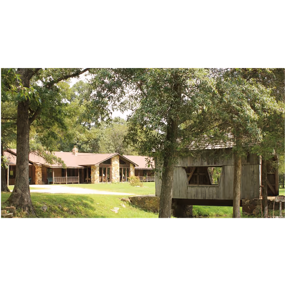 Single-story stone-front building with a covered porch on a grassy lawn surrounded by trees, with a rustic wooden covered bridge in the foreground.