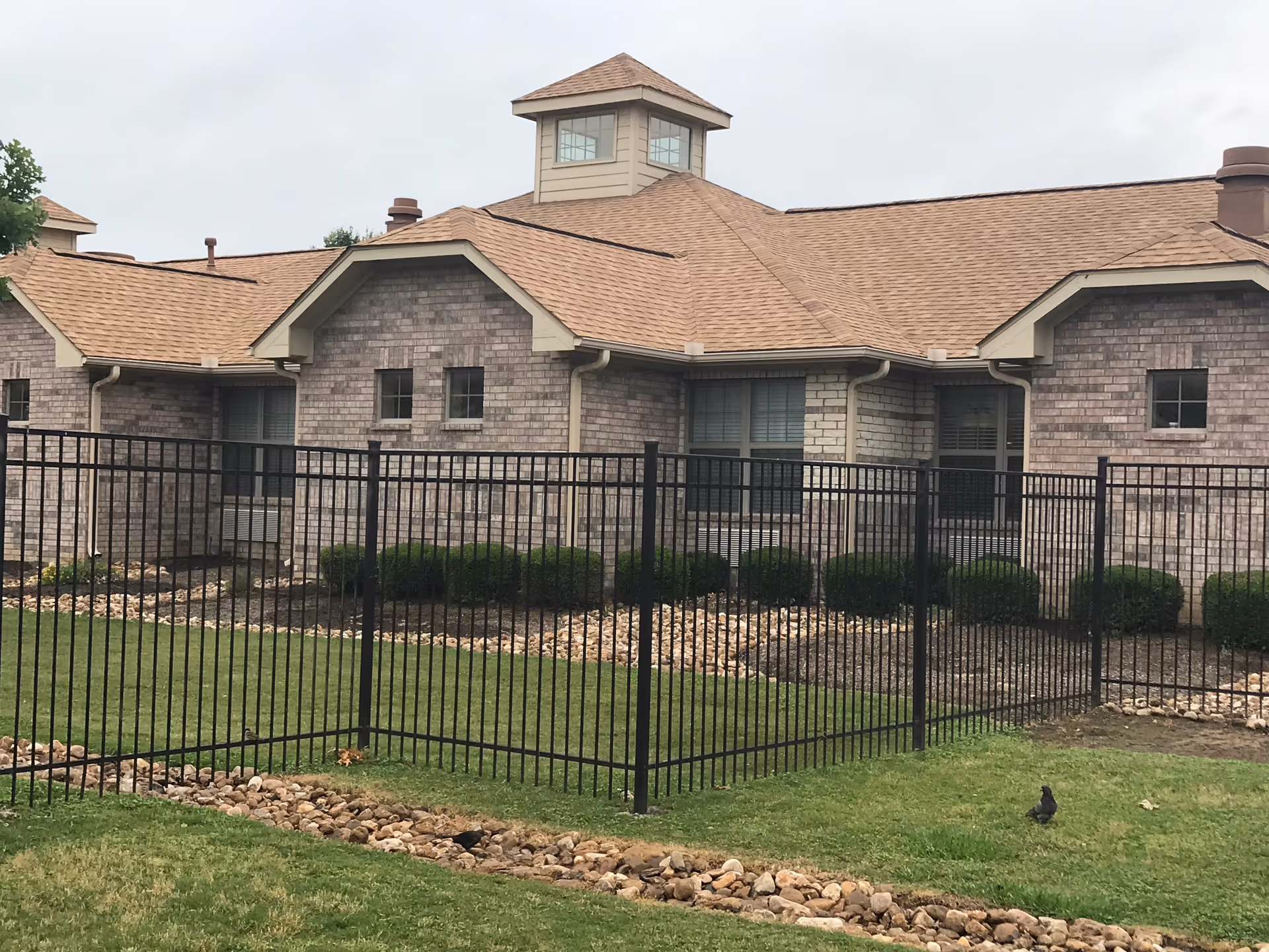 Brick single-story building with a tan roof, black metal fence, lawn, shrubs and rock landscaping in front.