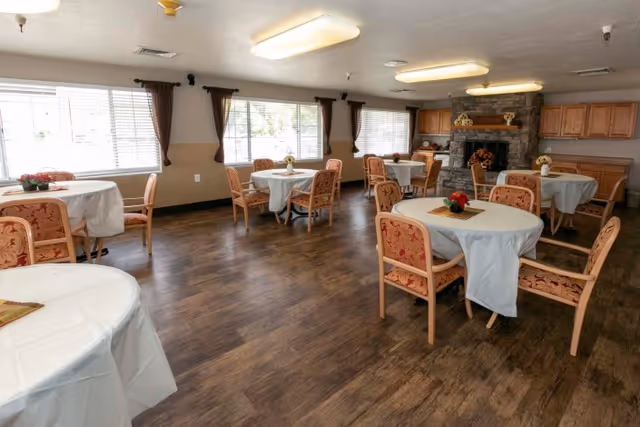 Spacious dining room with round tables draped in white tablecloths, upholstered chairs, large windows, and a stone fireplace.