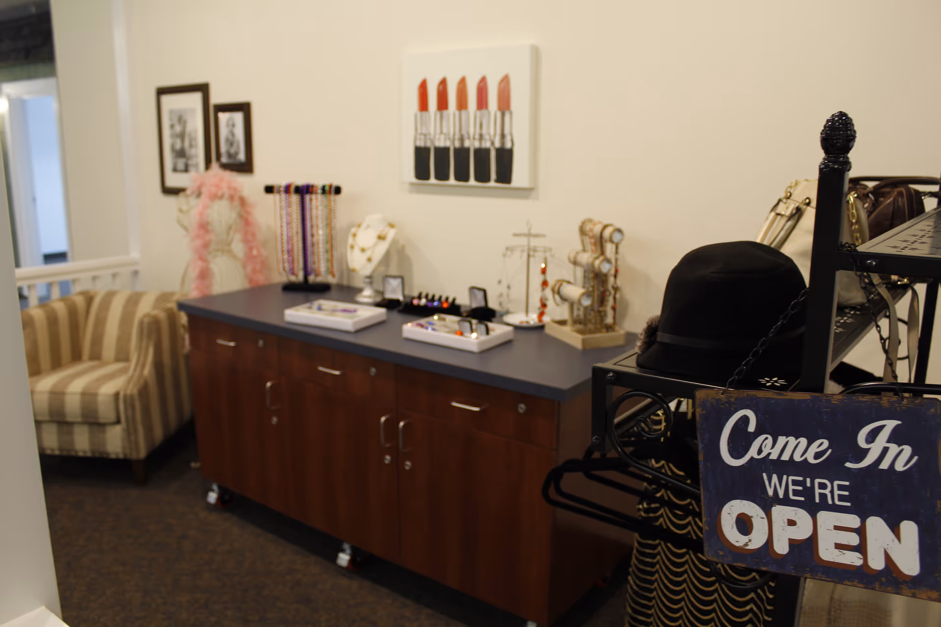 Interior view of a room with a wooden cabinet topped with jewelry displays including necklaces, bracelets, and rings. A striped armchair is visible in the background, along with framed pictures on the wall and a painting of lipsticks. In the foreground, a black hat is displayed on a rack with a sign that reads 'Come In We're OPEN'.