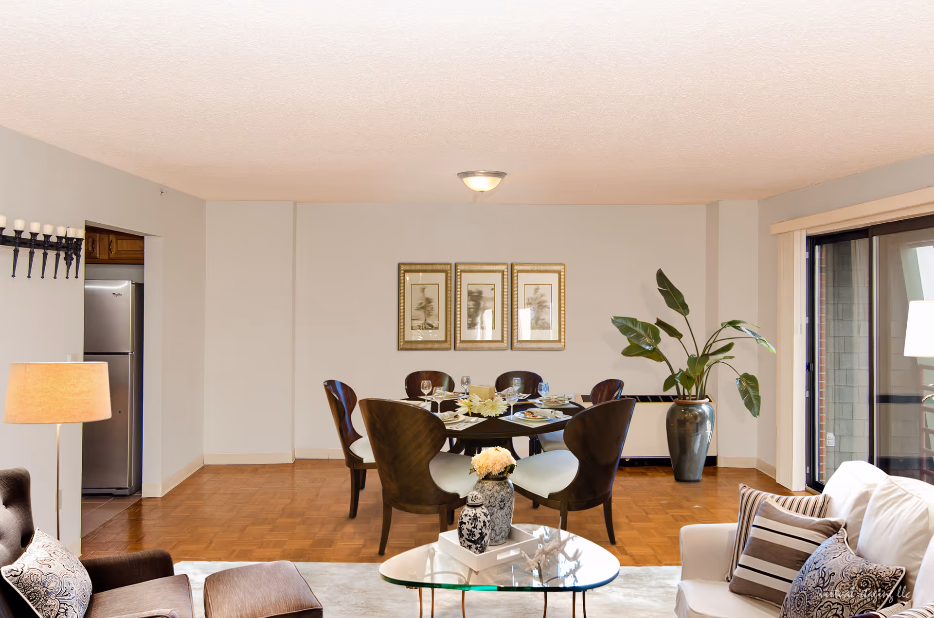 Bright open-plan living and dining area with a round dining table and chairs, sofa, glass coffee table and a large potted plant by sliding glass doors.