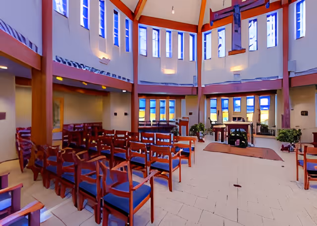 Interior view of a chapel or worship area with rows of wooden chairs with blue cushions arranged facing an altar. The space has tall windows with blue stained glass, high ceilings with wooden beams, and plants near the altar area.