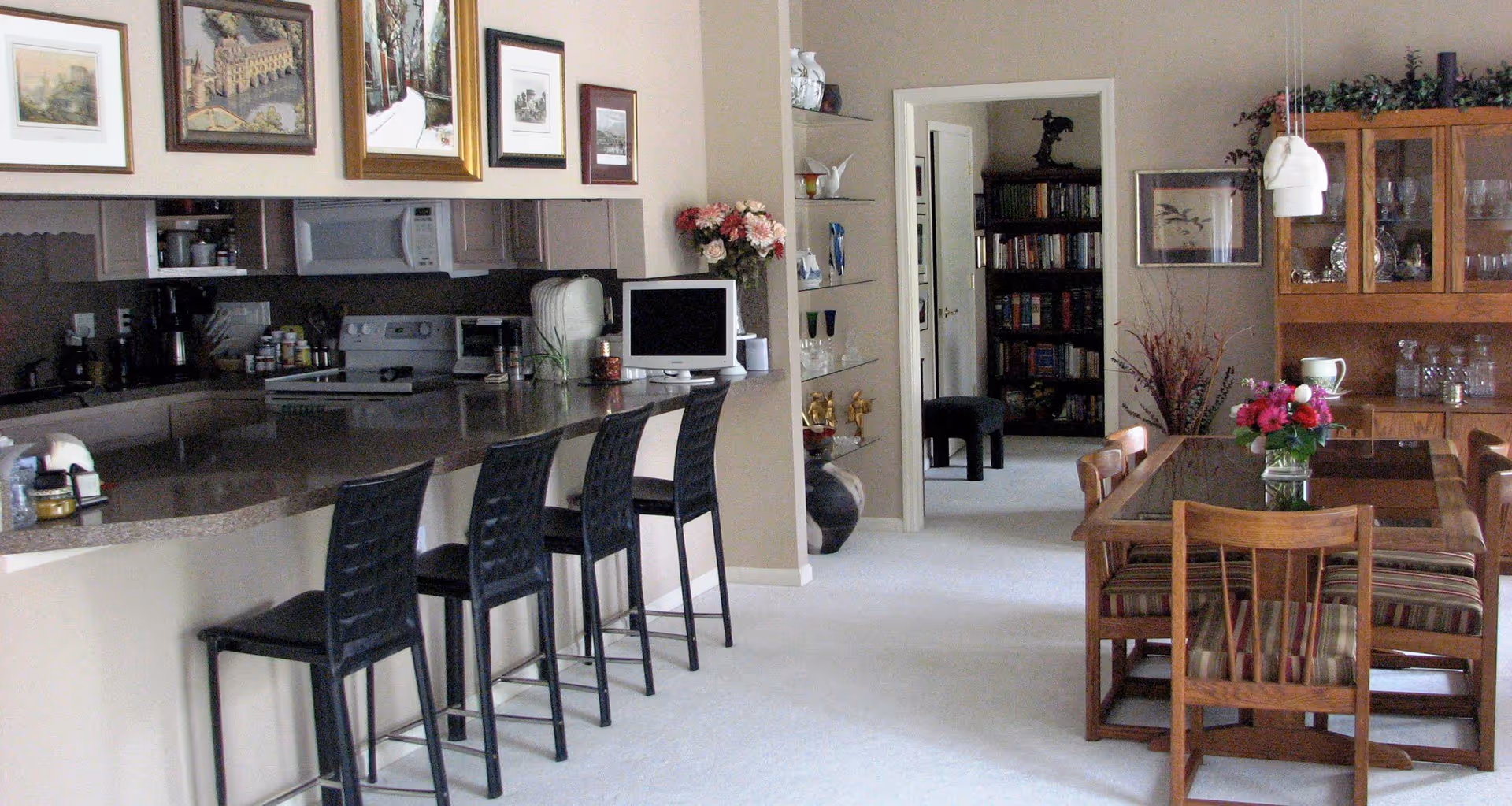 Open-plan interior showing a kitchen counter with four bar stools on the left and a wooden dining table with chairs and a vase of flowers on the right.