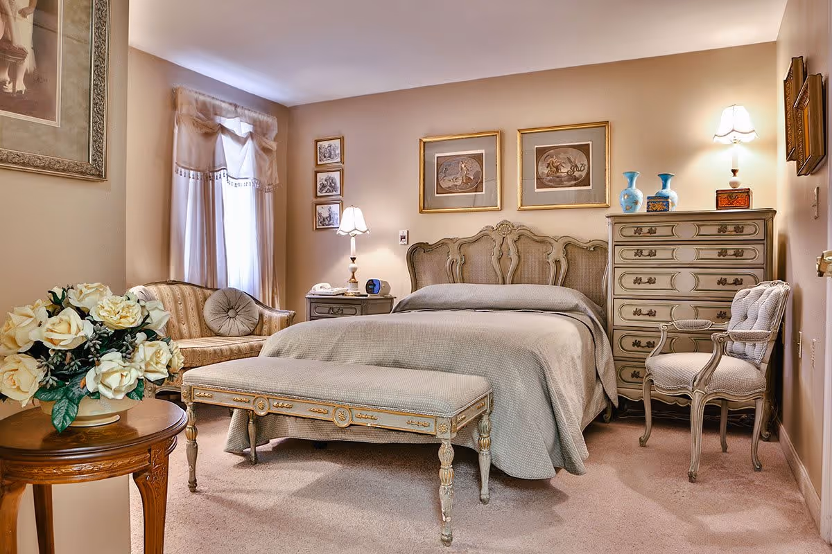 A cozy bedroom with a vintage style featuring a large bed with an ornate headboard and a matching bench at the foot. There is a small sofa near a window with sheer curtains, a wooden side table with a lamp and clock, a tall dresser with decorative vases, and an upholstered chair. The walls are adorned with framed artwork and the room is softly lit by lamps.