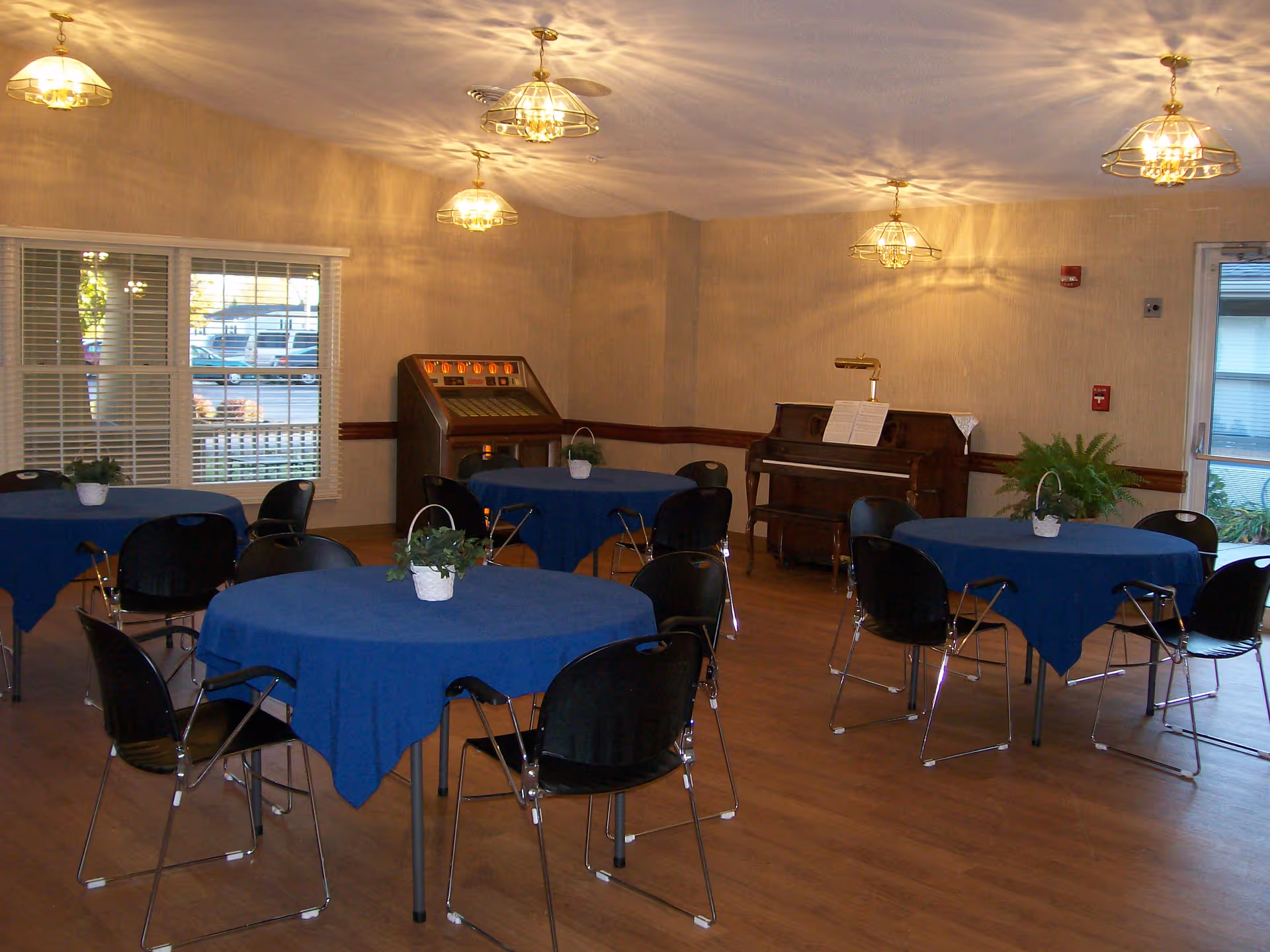 A communal dining/activity room with round tables covered in blue tablecloths, black chairs, a piano and a jukebox.