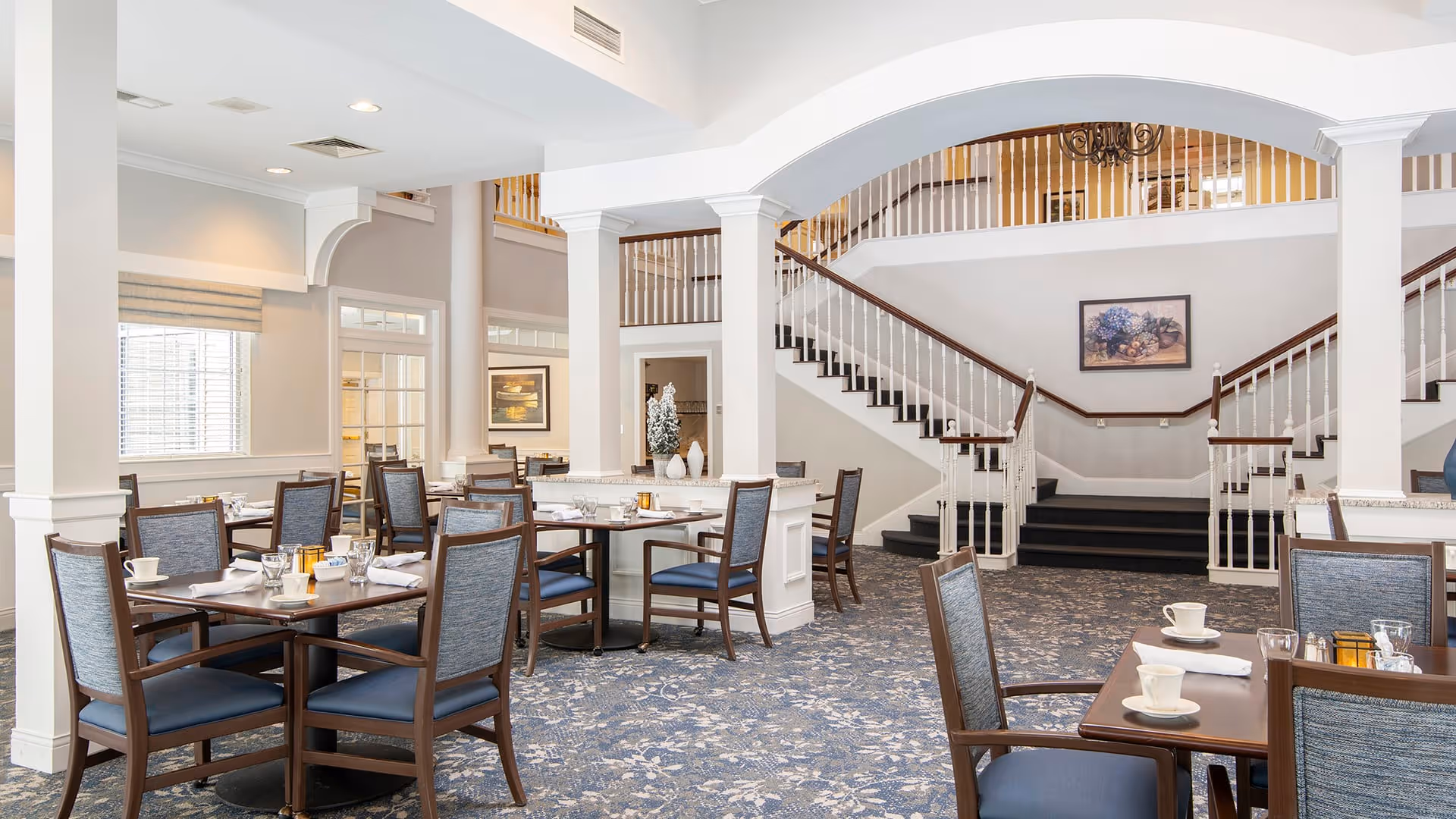 A spacious dining room with multiple wooden tables and chairs arranged neatly. Each table is set with white cups, glasses, and napkins. The room features a patterned carpet, white walls, large windows with blinds, and a staircase with wooden handrails leading to an upper floor. There is a framed picture on the wall above the staircase and decorative vases on a central counter.