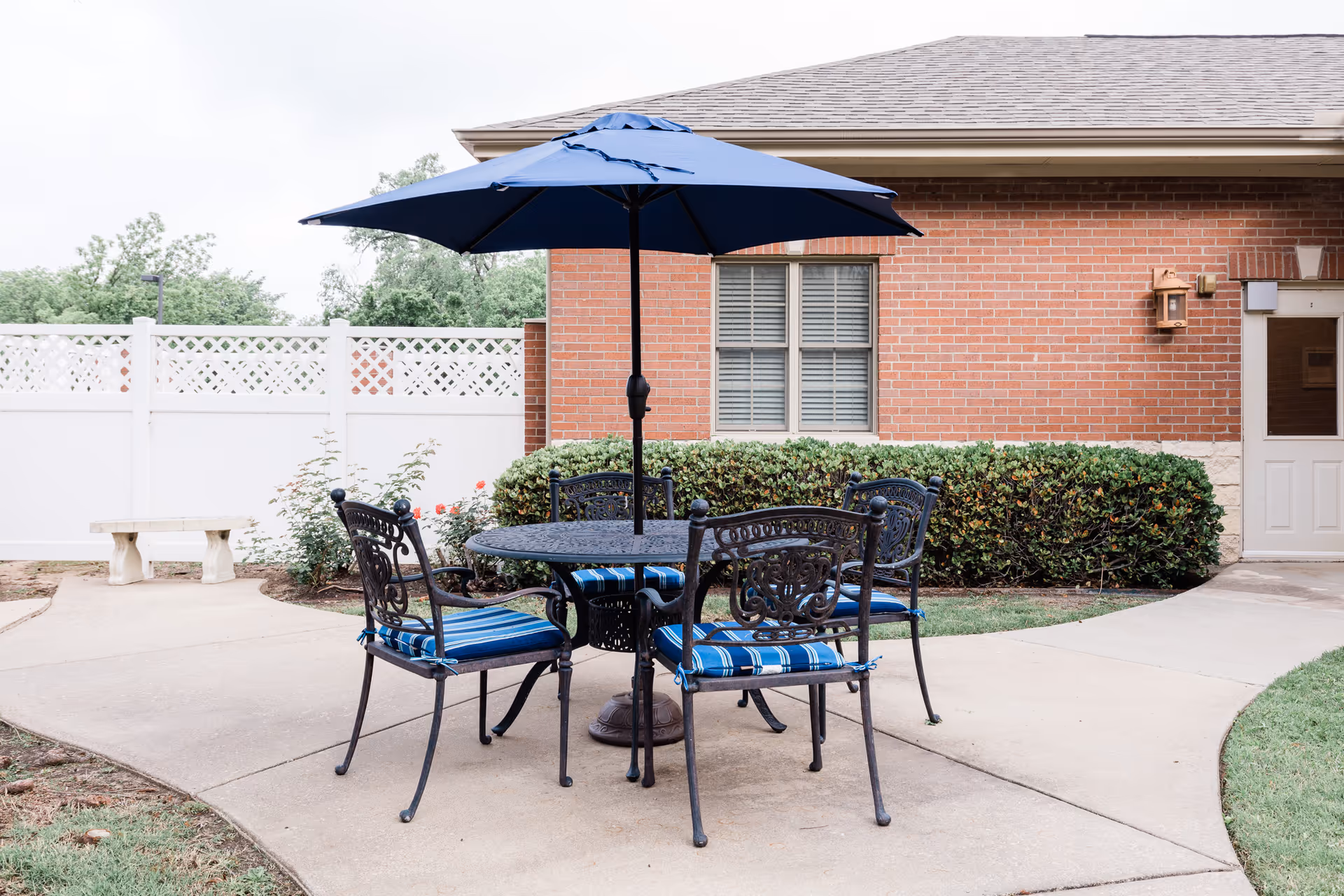 Outdoor patio area with a round black metal table and four matching chairs with blue striped cushions. A large blue umbrella is open above the table. The patio is paved and surrounded by green bushes, a white fence, and a brick building with a window and a door.