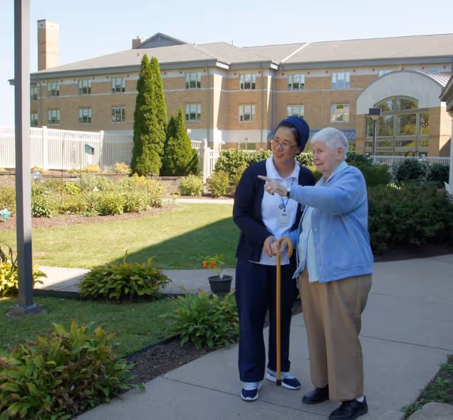 An elderly woman with a cane is standing outside on a paved pathway in a garden area, pointing at something in the distance. She is accompanied by a caregiver or nurse dressed in a navy blue uniform and head covering. Behind them is a large multi-story brick building with several windows, surrounded by green shrubs and trees.