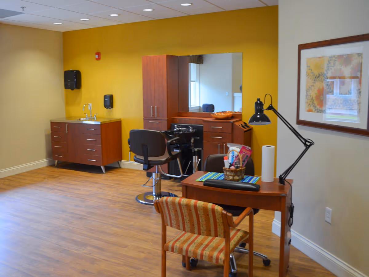 Interior room with wooden flooring and yellow accent wall featuring a salon station with a large mirror, a black salon chair, and wooden cabinets. There is a small desk with a striped chair, a desk lamp, and a basket with various items. A framed floral artwork hangs on the adjacent beige wall.