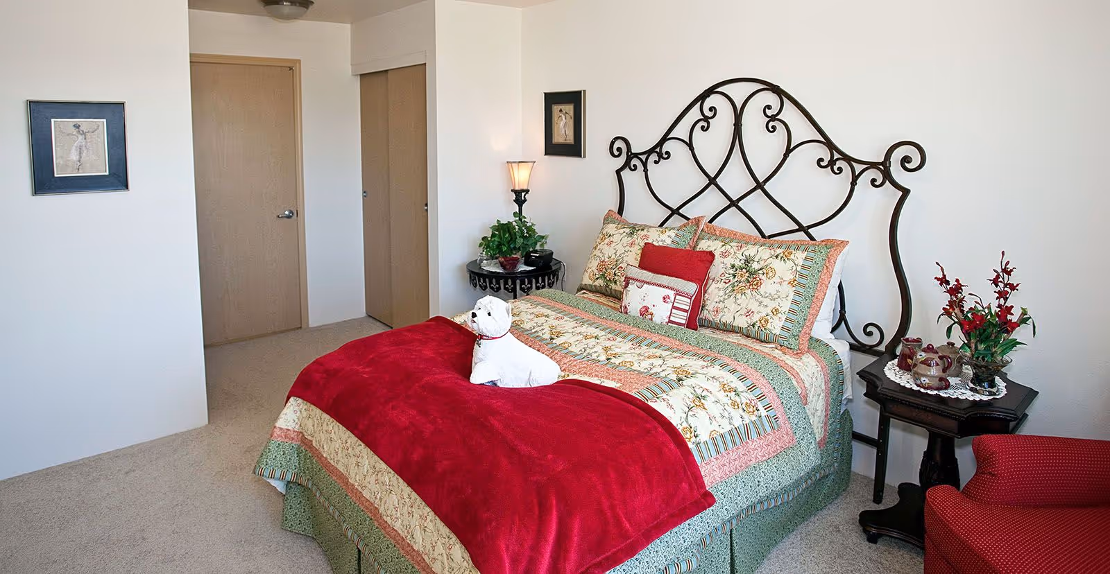 A cozy bedroom with a decorative wrought iron headboard, a bed covered with a floral and striped quilt, and a red blanket. A small white dog figurine is placed on the bed. There are two framed pictures on the walls, a round side table with a lamp and a plant, and another small table with a tea set and a flower arrangement. A red armchair is partially visible on the right side.