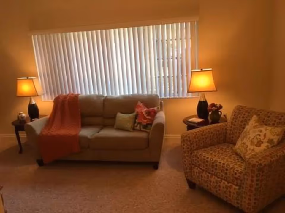 Cozy living room with a sofa, patterned armchair, side tables with lamps, and a large window with vertical blinds.