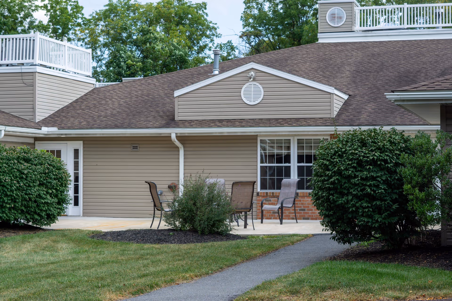 Outdoor patio area at Celebration Villa of Exeter with several chairs around a small table, surrounded by bushes and greenery, adjacent to a beige building with white trim and a brown shingled roof.