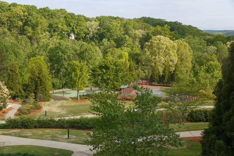 A scenic outdoor view of a senior living facility's landscaped grounds featuring lush green trees, manicured lawns, walking paths, and tennis courts surrounded by dense forest.