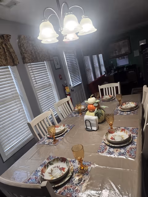 A dining room table set for six with autumn-themed plates and amber-colored glasses. The table has a beige tablecloth with a clear plastic cover and decorative placemats. A centerpiece made of stacked pumpkins is in the middle of the table. The room has several windows with white blinds and patterned valances, and a chandelier with five lights hangs above the table. In the background, there is a door and a living area with a red couch and a television.