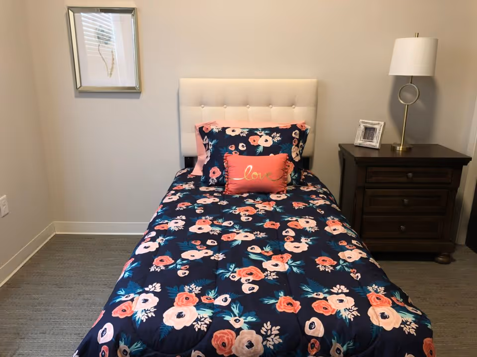 A neatly made single bed with a white tufted headboard, covered with a dark blue floral comforter featuring pink and peach flowers. There are matching pillows and a small rectangular pillow with the word 'love' embroidered on it. To the right of the bed is a dark wooden nightstand with two drawers, a small framed photo, and a modern lamp with a white shade. A framed botanical print hangs on the beige wall to the left of the bed.