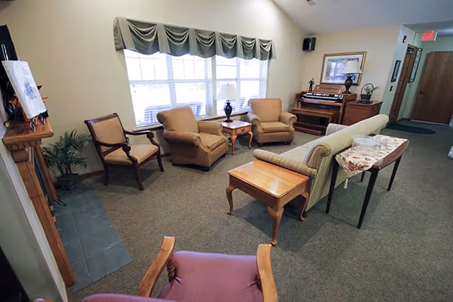 A cozy living room area in a senior living facility with several armchairs, a sofa, side tables, a lamp, a piano, and a window with a valance. The room has carpeted flooring and light-colored walls.