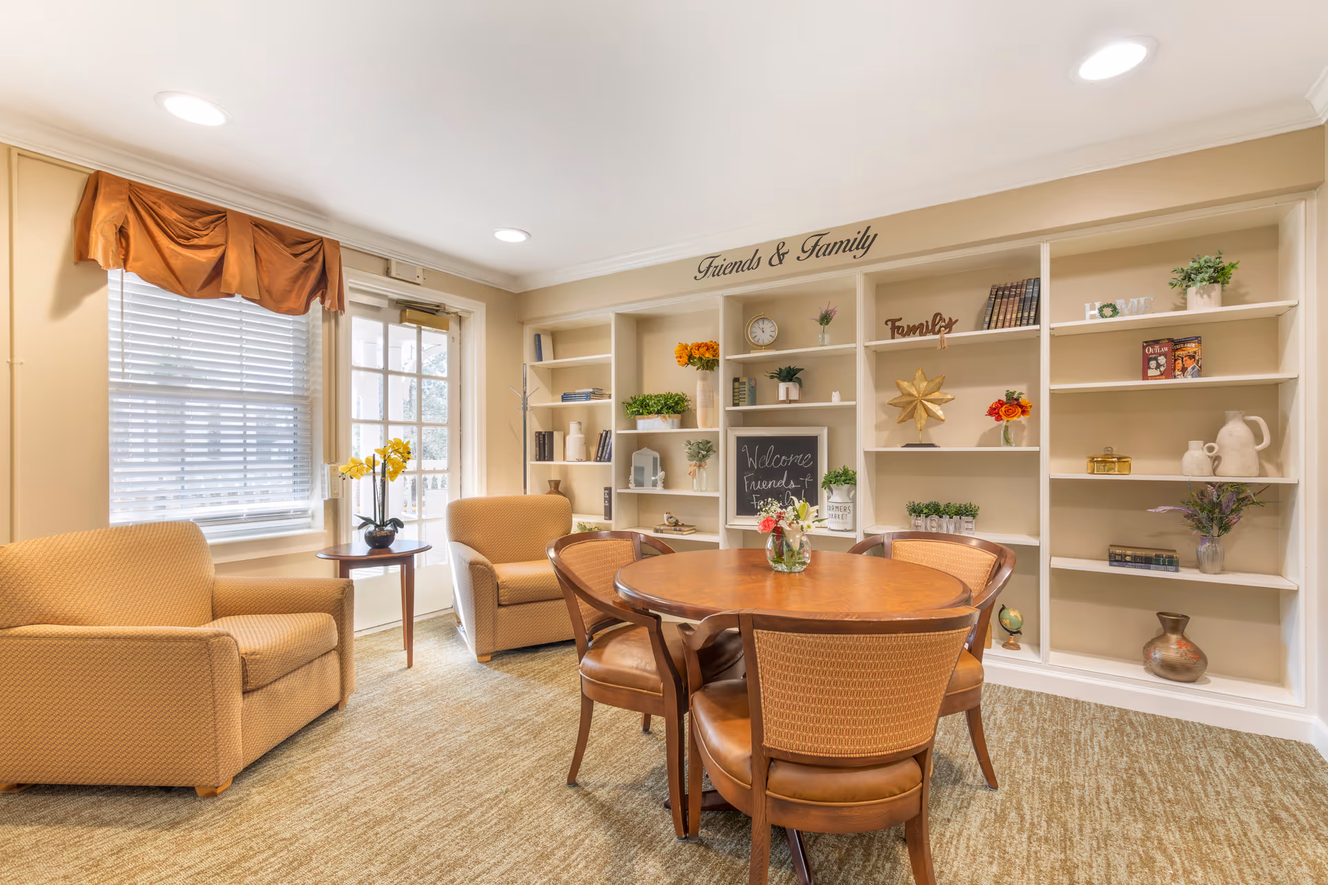 A cozy living room area with two beige armchairs, a round wooden table with four chairs, and built-in shelves decorated with books, plants, and decorative items. A window with blinds and a brown valance is on the left, and a door with glass panels is next to it. The shelves have words like 'Friends & Family' and 'Family' displayed.