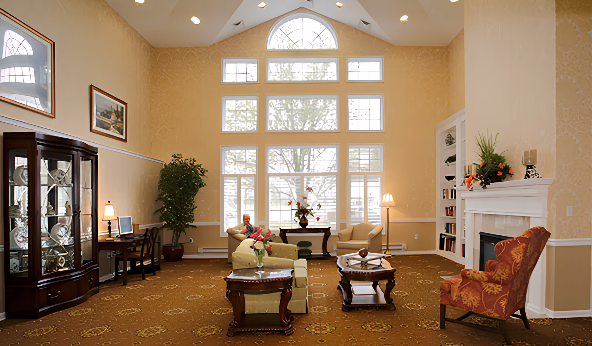 A spacious living room with high vaulted ceilings and large windows letting in natural light. The room features a beige sofa, a red patterned armchair, two wooden coffee tables, a fireplace with decorative plants on the mantel, a tall potted plant, a wooden display cabinet with glass doors, a desk with a lamp and computer, and framed artwork on the walls.