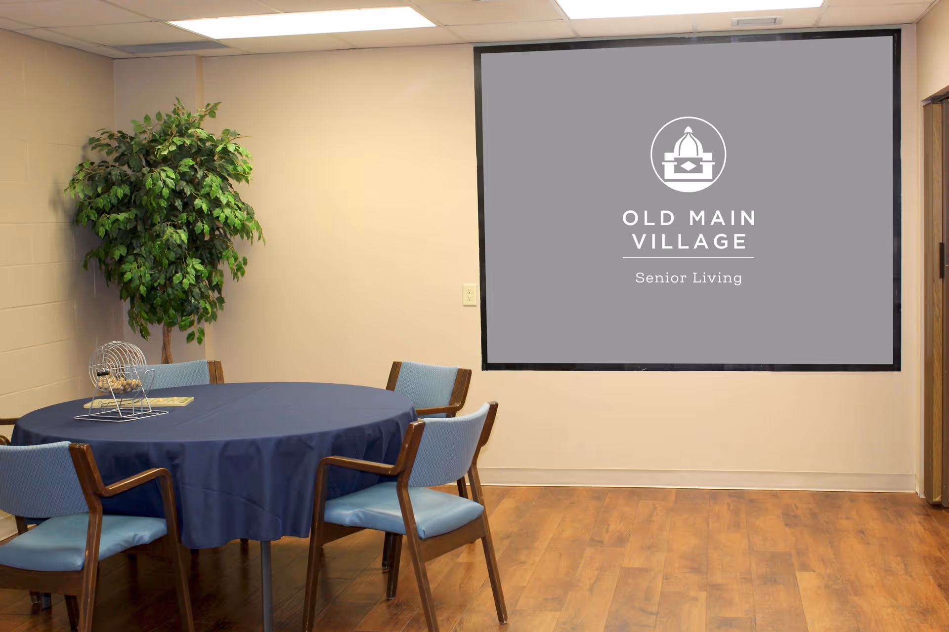 A small meeting or activity room with a round table covered by a blue tablecloth and surrounded by four blue cushioned chairs with wooden arms. There is a bingo cage with balls on the table and a potted artificial tree in the corner. On the wall is a large screen or board displaying the Old Main Village Senior Living logo.