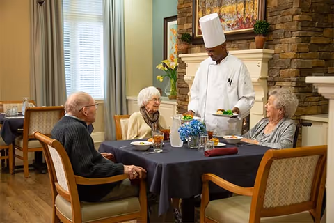 A chef serves meals to three elderly residents seated at a table in a cozy assisted-living dining room.