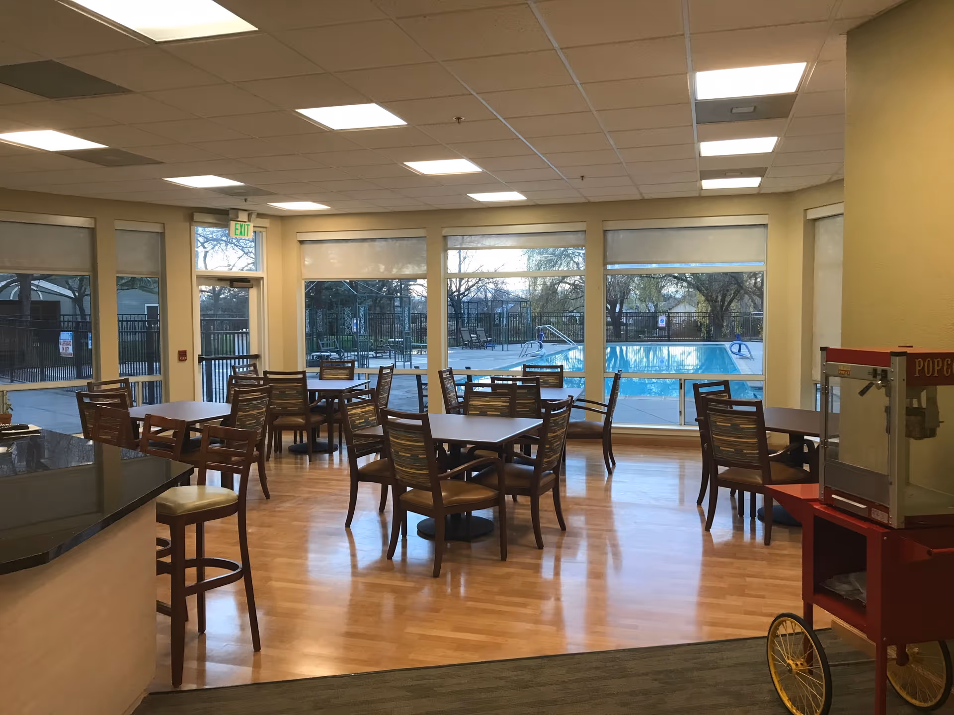 Dining area with several tables and chairs arranged on a wooden floor, large windows showing an outdoor swimming pool and trees, and a popcorn machine on the right side.