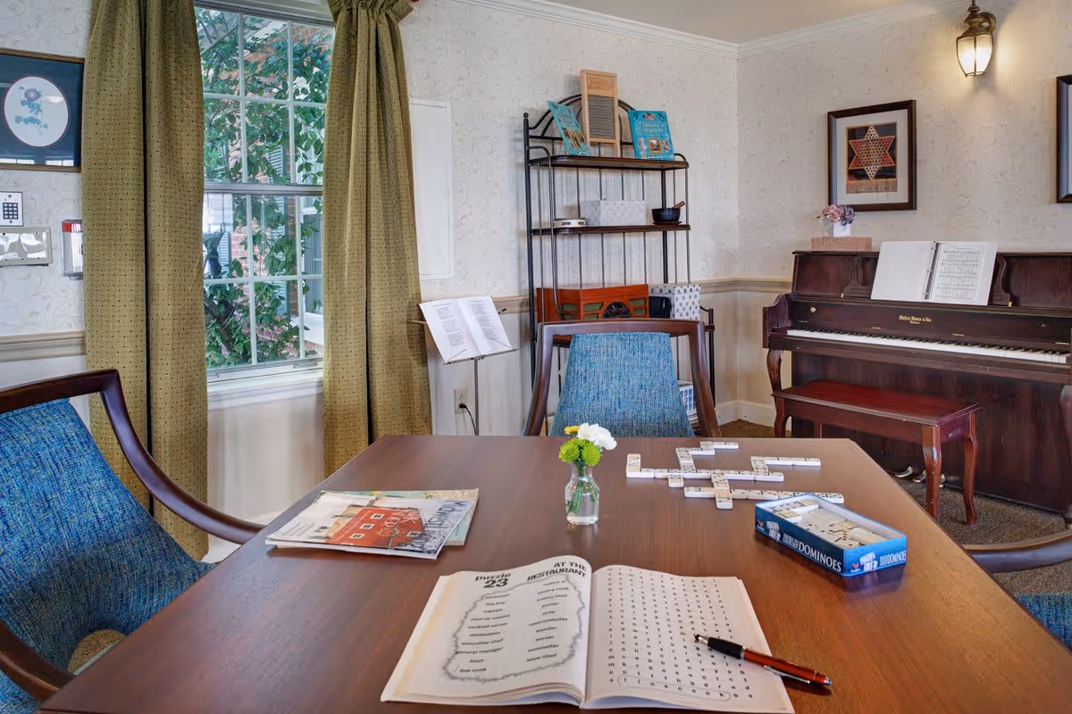A cozy room with a wooden table featuring a word search puzzle book, a pen, a small vase with flowers, and a game of dominoes in progress. The room has blue cushioned chairs, a window with green curtains, a piano with sheet music, framed artwork on the walls, and a black metal shelf with books and decorative items.