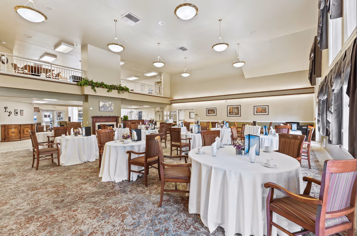A spacious dining room with round tables covered in white tablecloths, each set with napkins, cups, and small flower arrangements. Wooden chairs surround the tables. The room has high ceilings with multiple hanging light fixtures, large windows with dark curtains, and a carpeted floor. There is a mezzanine level visible in the background with additional seating and a decorative plant on the ledge.