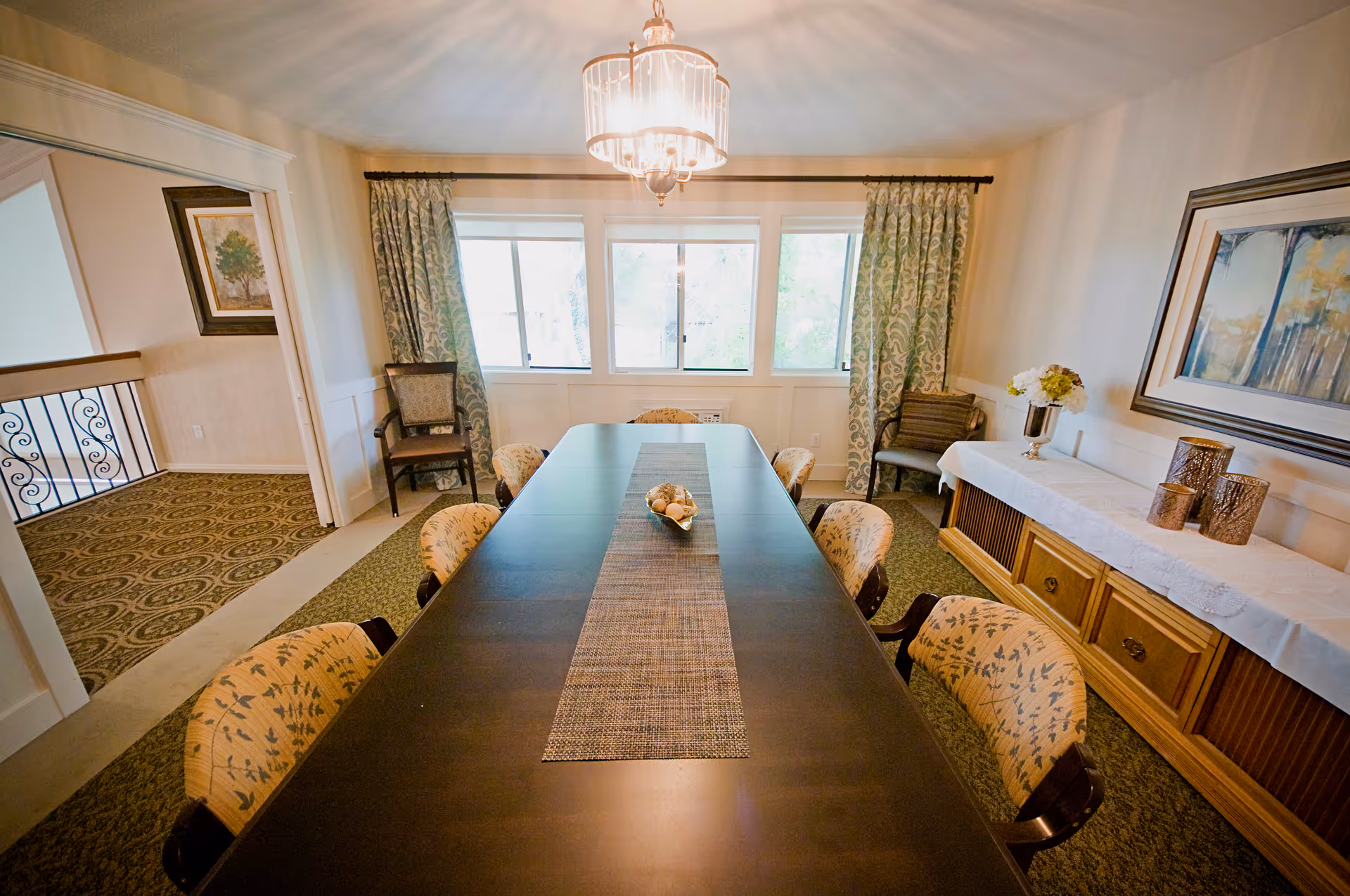 A well-lit dining room with a long dark wooden table surrounded by eight upholstered chairs with floral patterns. The room features large windows with patterned curtains, a chandelier hanging above the table, a sideboard with decorative items and flowers, and framed artwork on the walls.