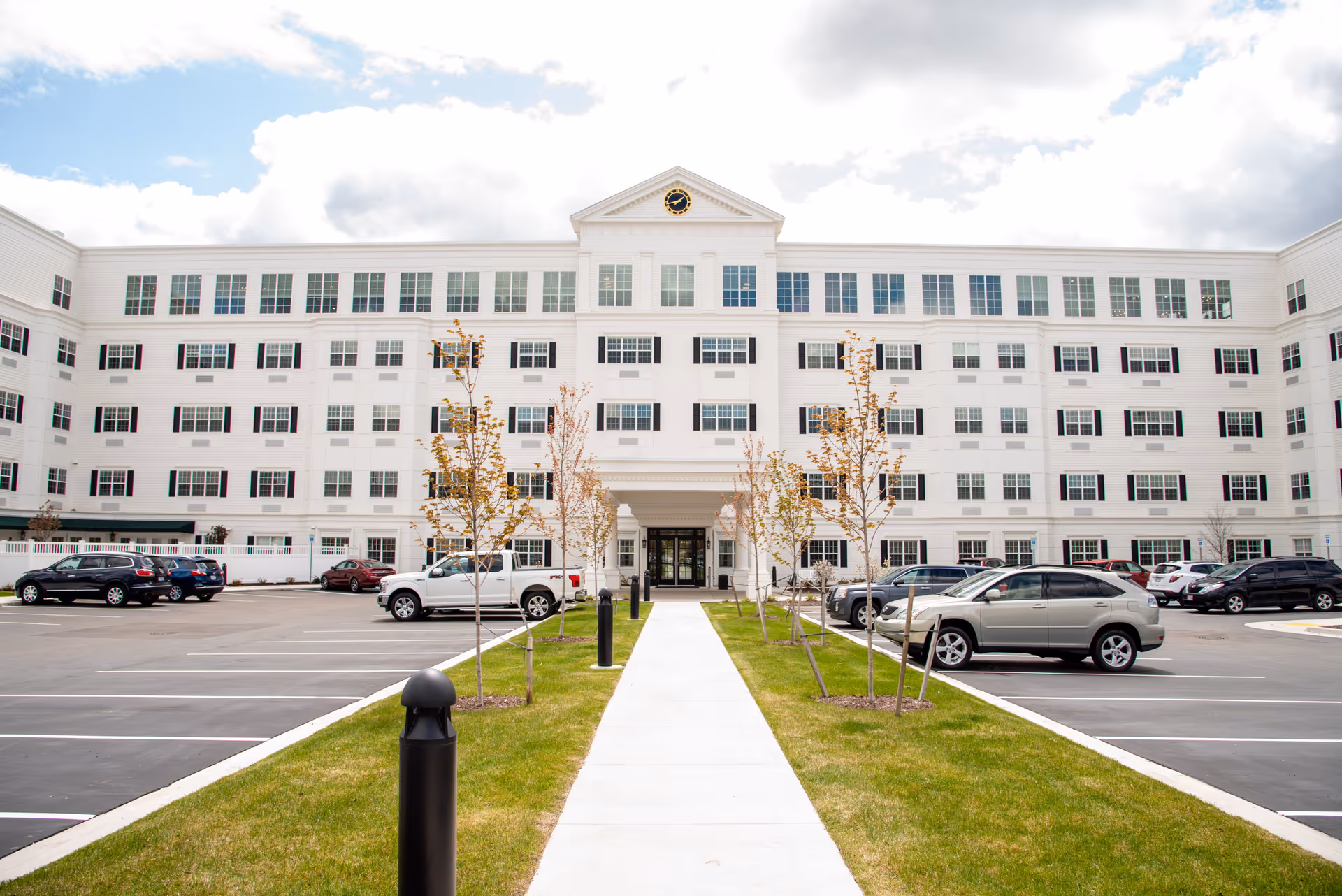 Front exterior of a large white multi-story senior living building with a central entrance, parking lot, and walkway.