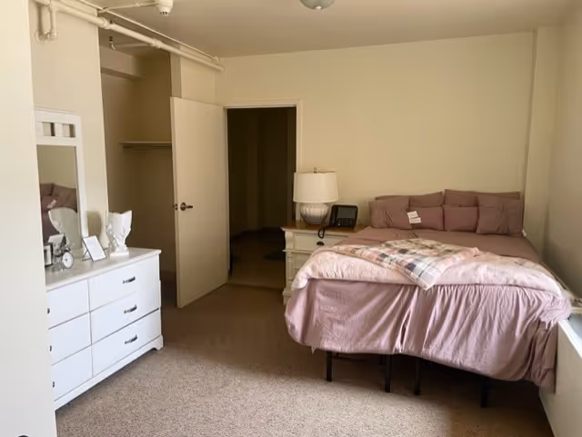 A bedroom with a bed covered in pink bedding and multiple pillows, a white nightstand with a lamp and a phone, a white dresser with a mirror and decorative items, and an open door leading to a closet and another room.