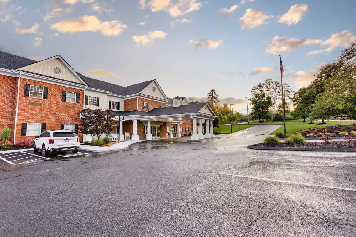 Exterior view of a two-story senior living facility building with a brick and white facade, a covered entrance with columns, a parked white SUV, an American flag on a flagpole, and landscaped greenery under a partly cloudy sky.