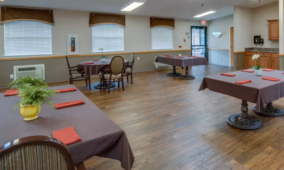 A dining room with several tables covered in brown tablecloths, each set with red napkins and small potted plants. The room has wooden flooring, three windows with blinds and valances, and a door leading outside. There are chairs around the tables and a small kitchenette area with cabinets and a coffee maker in the corner.