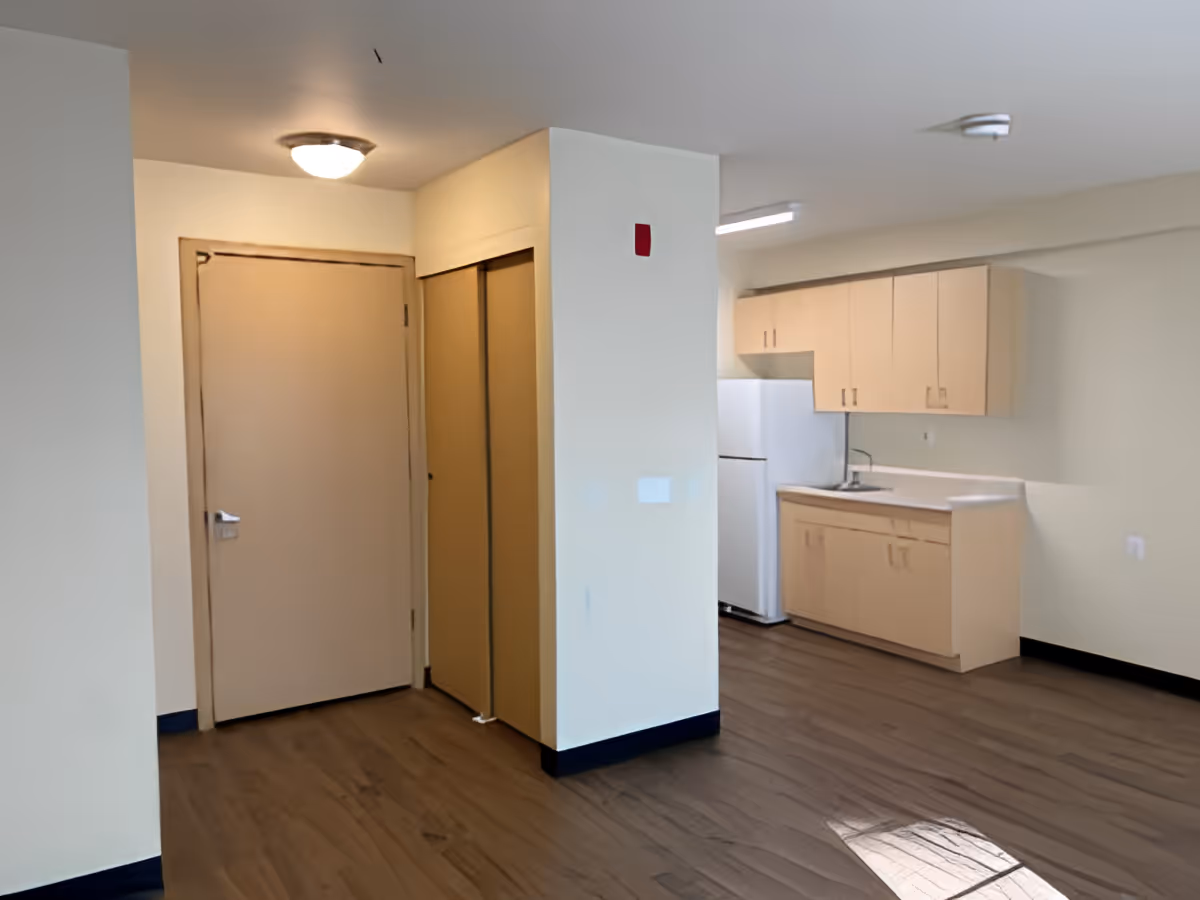 Interior view of a small kitchen area with light wood cabinets, a white refrigerator, and a sink. To the left, there is a closed door and a closet with sliding doors. The room has light-colored walls and wood flooring.