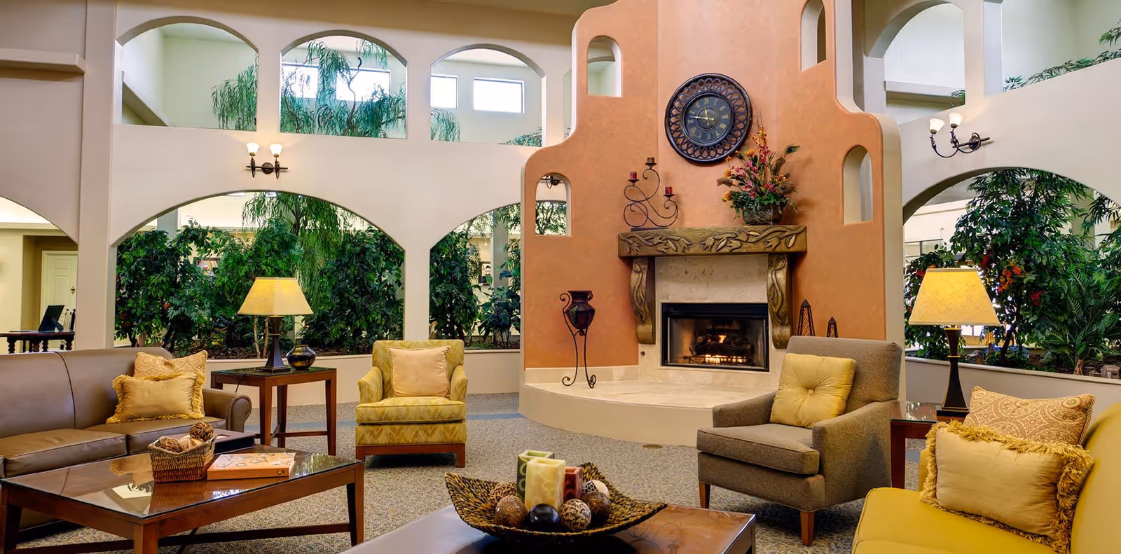 A cozy living room area in a senior living facility with a fireplace set in a decorative orange wall. The room features comfortable seating including a brown leather sofa, yellow and gray armchairs, and a wooden coffee table with decorative items. There are large indoor plants and arched windows allowing natural light to fill the space.