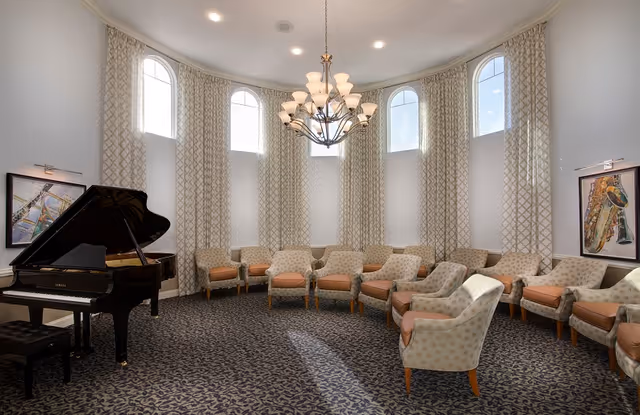 A bright, elegant room with a curved arrangement of cushioned armchairs around the perimeter. The room features tall arched windows with patterned curtains, a grand piano with a bench on the left side, two framed musical instrument paintings on the walls, and a large chandelier hanging from the ceiling.