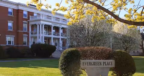 Brick multi-story building with white-columned front entrance and a lawn sign that reads 'Evergreen Place'.
