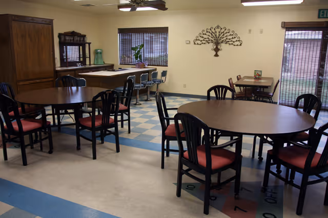 Interior view of a dining area in a senior living facility with multiple round wooden tables surrounded by black chairs with red cushions. In the background, there is a counter with bar stools, a wooden cabinet, a wall decoration, and windows with blinds.