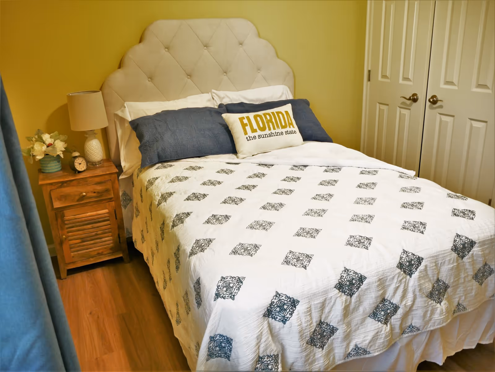 A cozy bedroom with a bed featuring a tufted beige headboard, white and blue bedding, and a decorative pillow that says 'FLORIDA the sunshine state'. There is a wooden nightstand with a lamp, a small clock, and a flower arrangement. The room has yellow walls, wooden flooring, and a closed double-door closet.