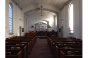Interior of a chapel with rows of wooden pews leading to a raised pulpit or altar beneath a decorative ceiling and chandelier.