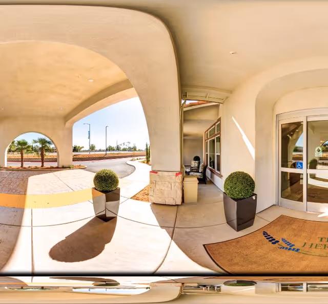 Covered entrance area of a senior living facility with two large potted plants, a doormat, and automatic sliding glass doors. The area is shaded by a large archway, and there is a pathway leading to the outside with palm trees and a clear sky visible in the background.