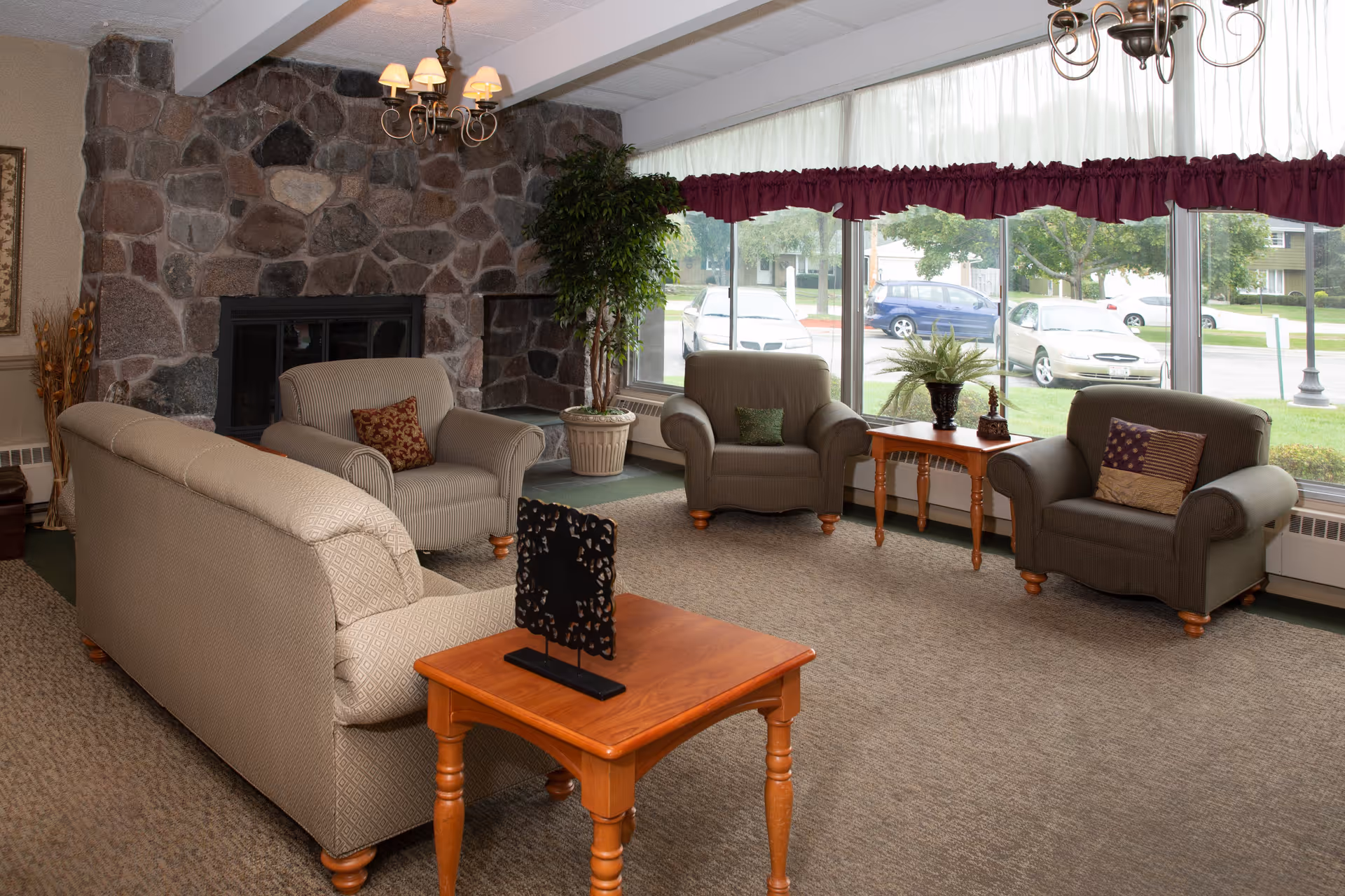 A cozy living room with beige and brown upholstered armchairs and a sofa arranged around a wooden coffee table. There is a stone fireplace on the left wall and large windows with burgundy valances letting in natural light. A potted plant and decorative items are placed on side tables near the windows.