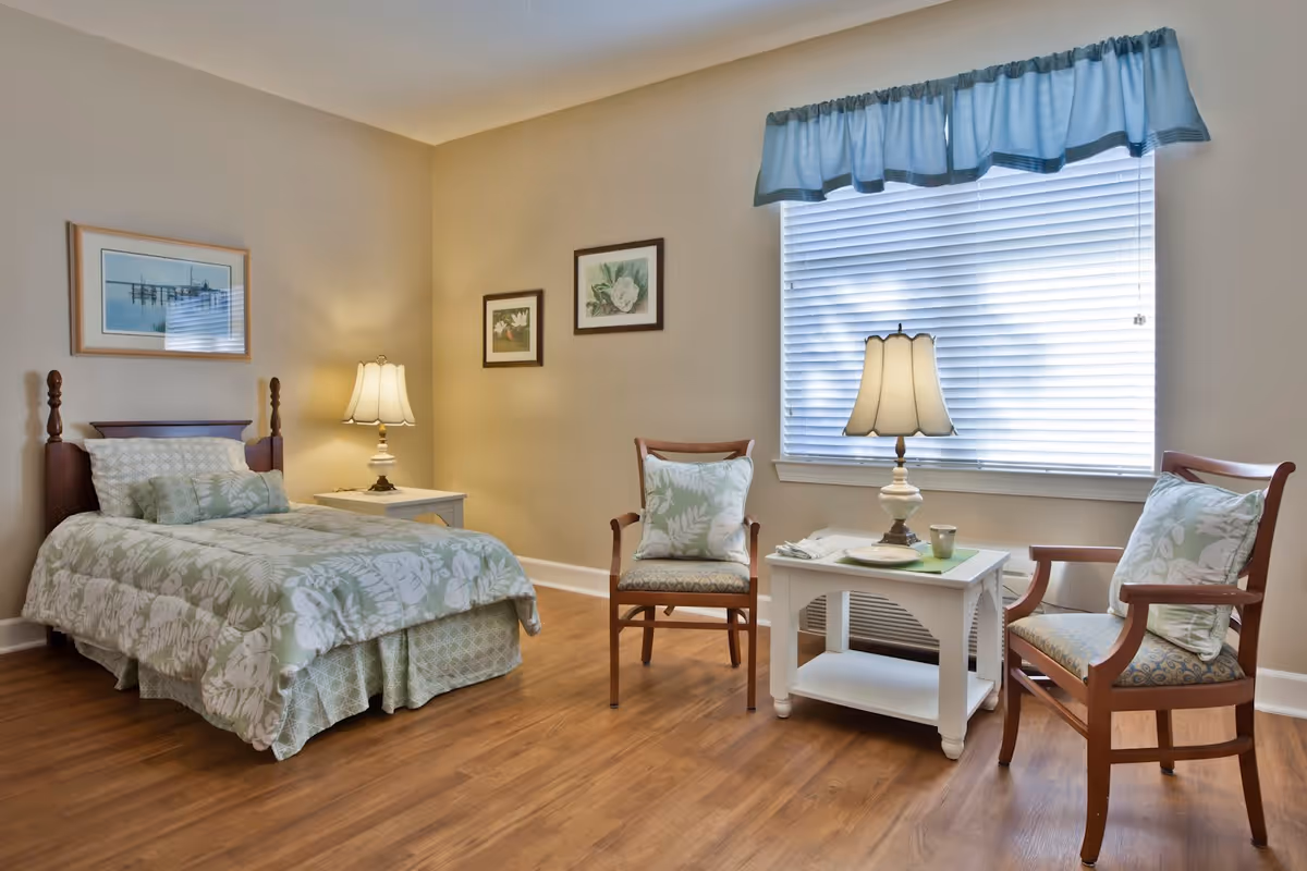 A cozy bedroom with a single bed covered in green and white floral bedding, a wooden headboard, and a bedside table with a lamp. Two wooden chairs with cushions and a small white table with a lamp and a cup are placed near a large window with blinds and a blue valance. The room has wooden flooring and light-colored walls with framed artwork.