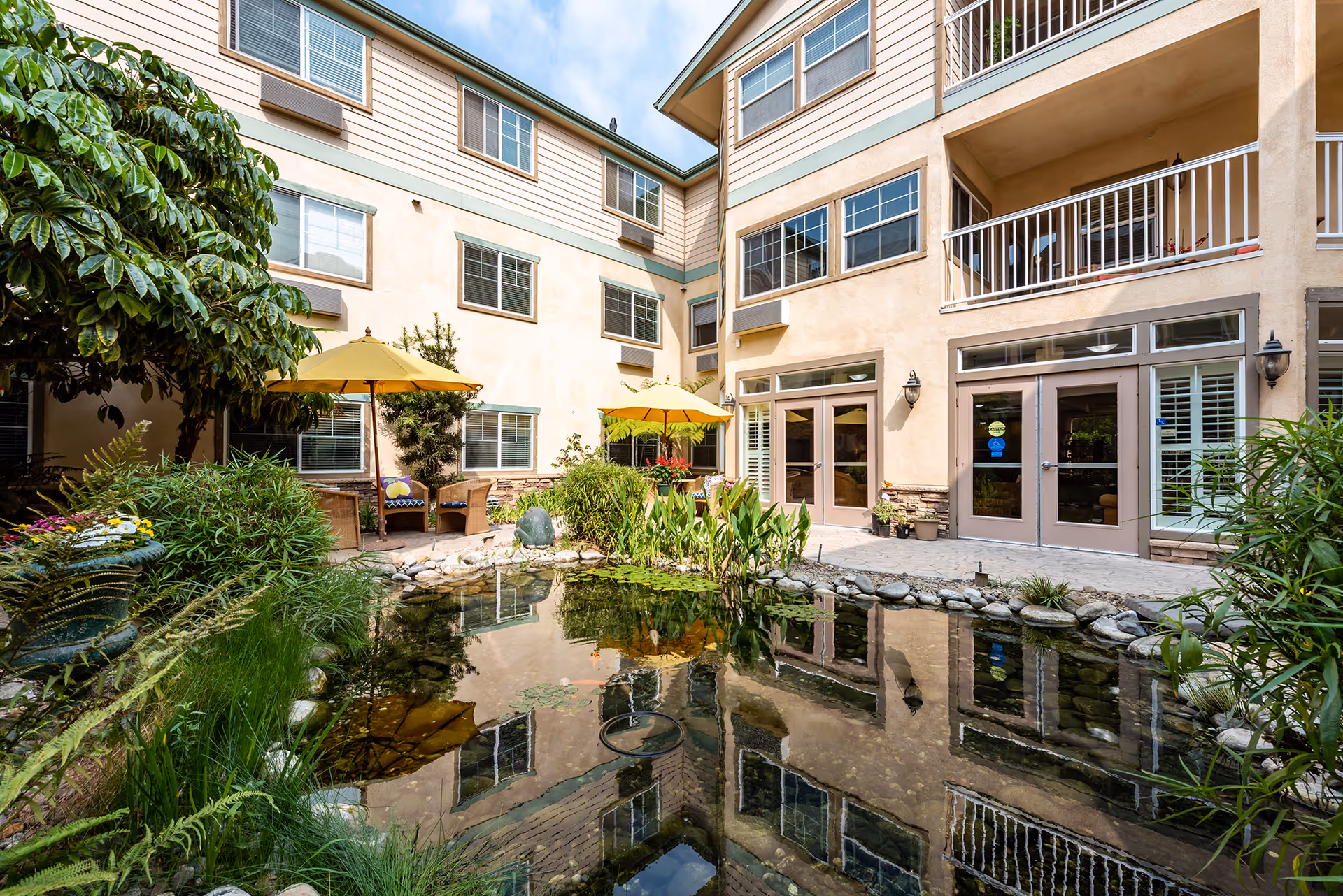 Outdoor courtyard area of a senior living facility with a small pond surrounded by rocks and plants. There are two yellow umbrellas shading wicker chairs and tables. The building has multiple windows and balconies overlooking the courtyard.